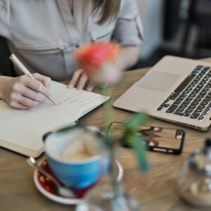 Woman writing in a notebook with a laptop and coffee cup on a desk. Ideal for workspace inspiration.